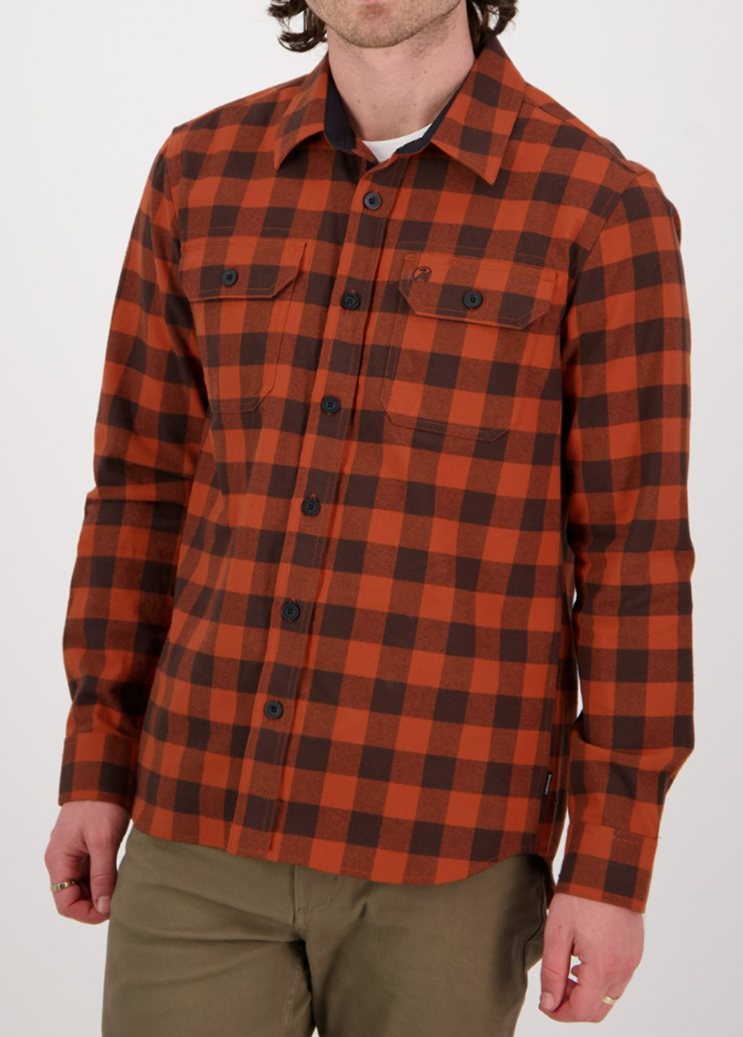 A person wearing the Swanndri Taranaki Tailor Shirt, a stylish orange and black plaid design with black buttons, crafted from 100% cotton, paired with olive green pants. This comfortable shirt features two chest pockets and long sleeves against a plain white backdrop.