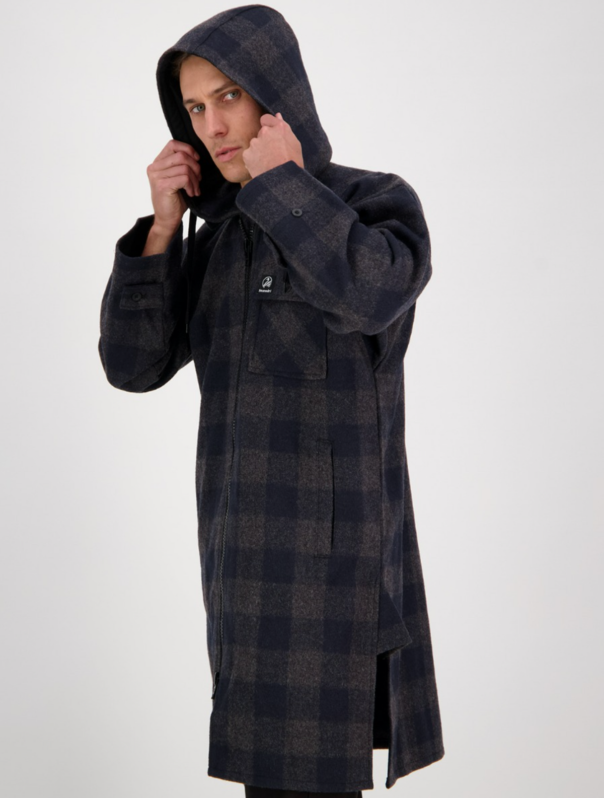 A person wearing the Swanndri Mosgiel Zip Front Bush Shirt, a long dark plaid coat with a detachable hood, looks to the side and holds up the hood with both hands against a plain white background.