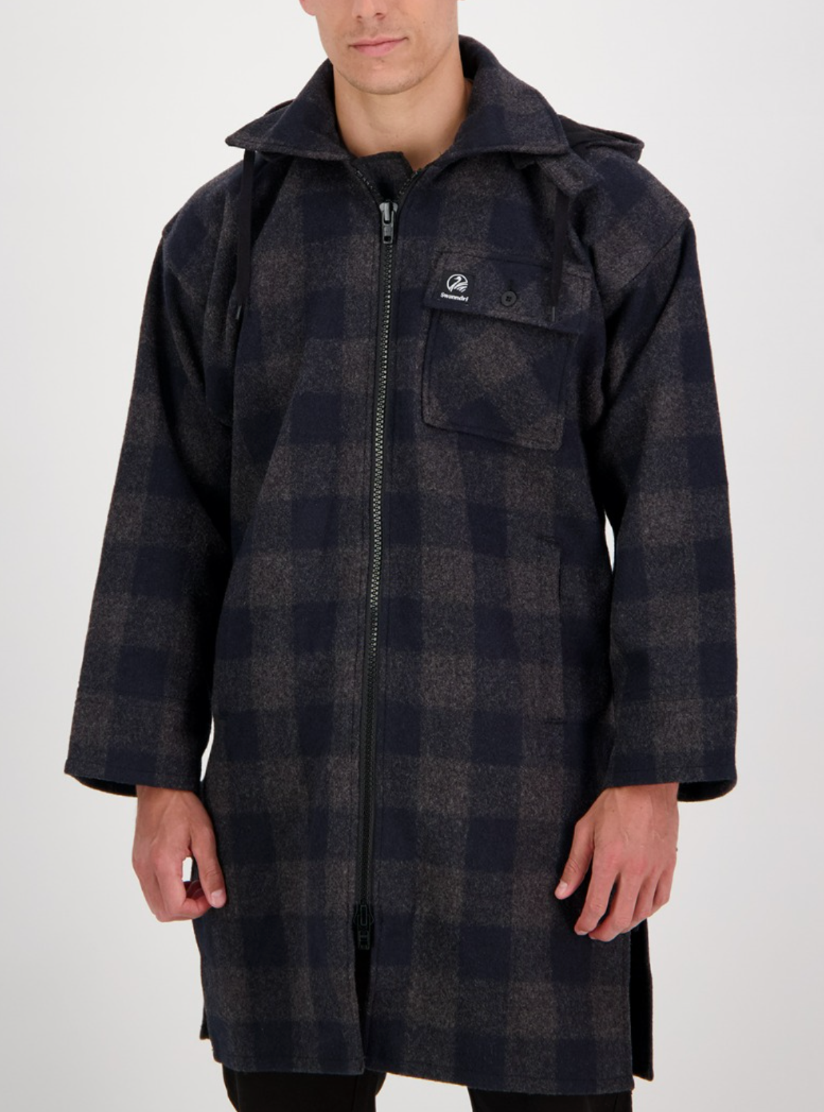 A person wearing the Swanndri Mosgiel Zip Front Bush Shirt—a long, dark plaid 100% wool overshirt with a zipper closure, button-flap chest pocket, and detachable hood—stands against a plain white background.