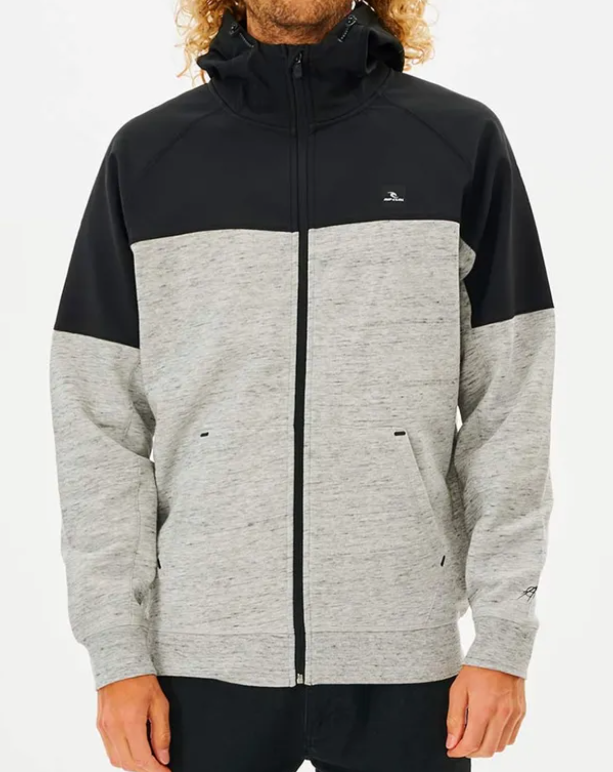 A person is wearing the Rip Curl Viral Anti Series Zip Thru Hood, which has a black top part and a light gray bottom part. This winter hoodie features a front zipper, side pockets, and small logos on the chest and left sleeve. The person, who has long curly hair, is standing against a plain white background.