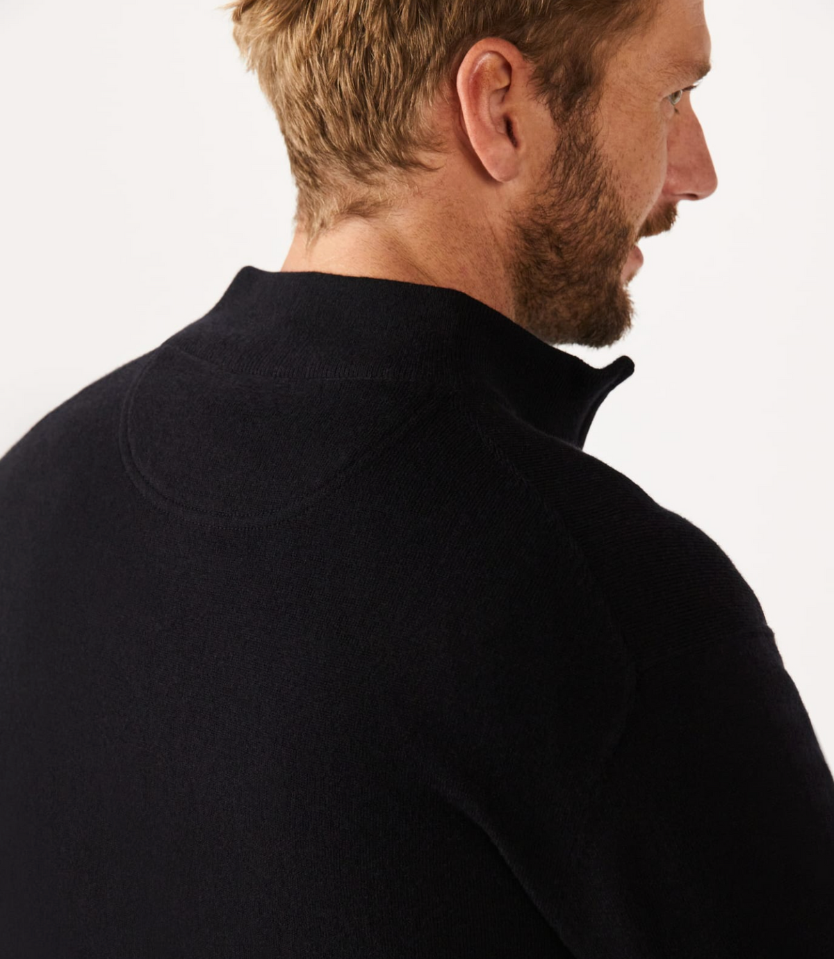 A man with short blonde hair and a beard, facing right, wears a dark-colored RM Williams Ernest Sweater. This luxurious 1/4 zip high-collared sweater is made of Merino wool against a plain white background.