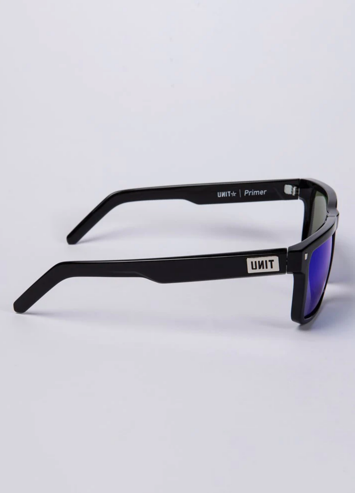 Side view of UNIT Primer Polarised Eyewear showcasing a rectangular, lightweight black frame. The straight arms feature the "UNIT" brand name, and the polarized lenses have a reflective blue tint with UV protection. The background is light gray.