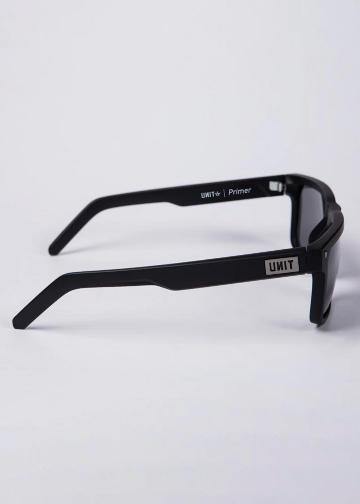 UNIT Primer Polarised Eyewear shown in black with a lightweight wraparound frame and thick arms. The UNIT logo and Primer model name are printed in white, providing UV protection and stylish design against a white background.