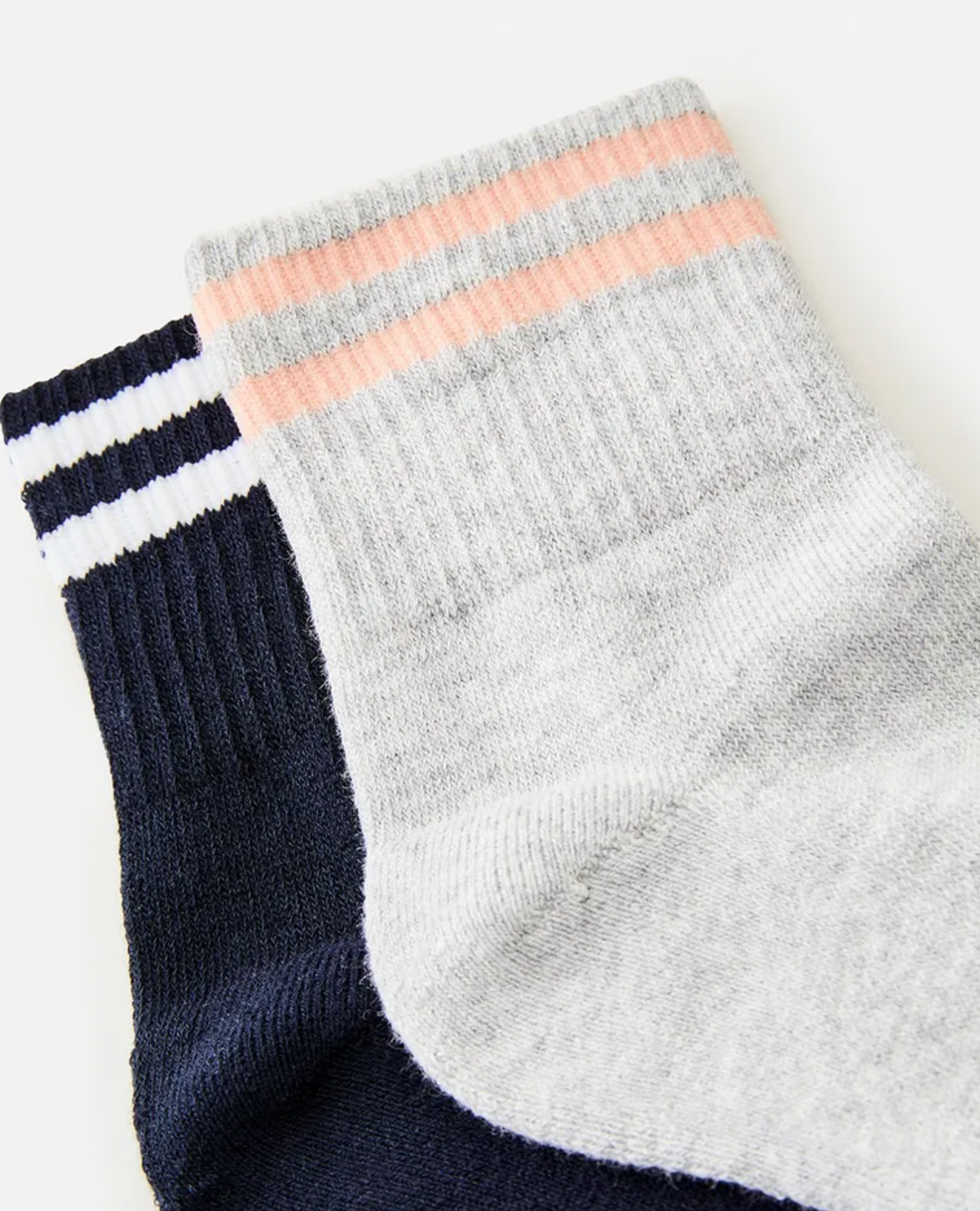 Close-up of two pairs of socks from the Rip Curl Womens VaporCool Sport Sock 2 Pack, showcasing their ribbed texture ideal for moisture-wicking comfort. The sock on top is light grey with peach and grey stripes at the cuff, while the sock underneath is dark blue with white stripes at the cuff.