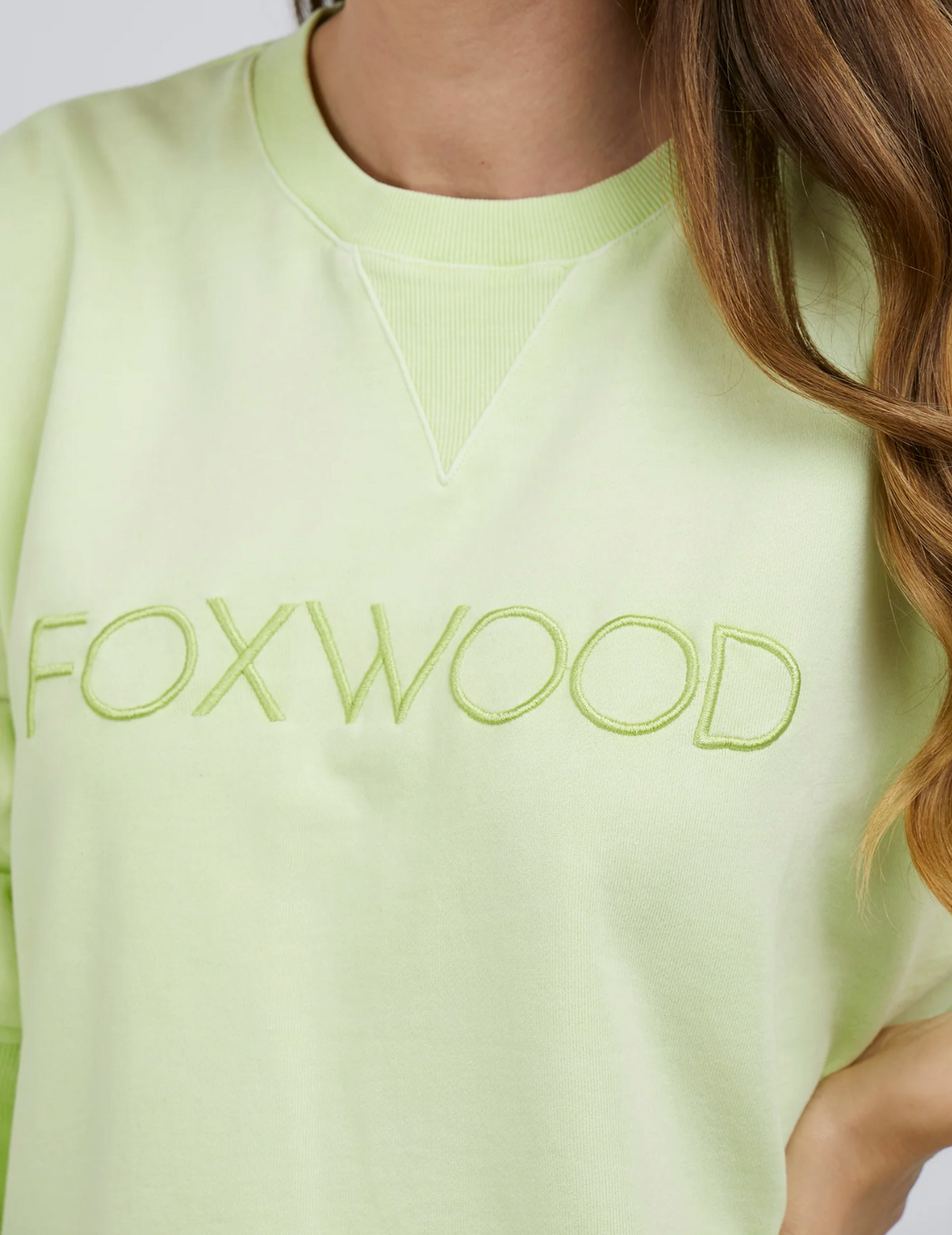 A close-up of a person in the Foxwood Simplified Crew, a light green cotton sweatshirt with Foxwood embroidery on the chest. The relaxed fit is paired with long, wavy brown hair draped over the right side.