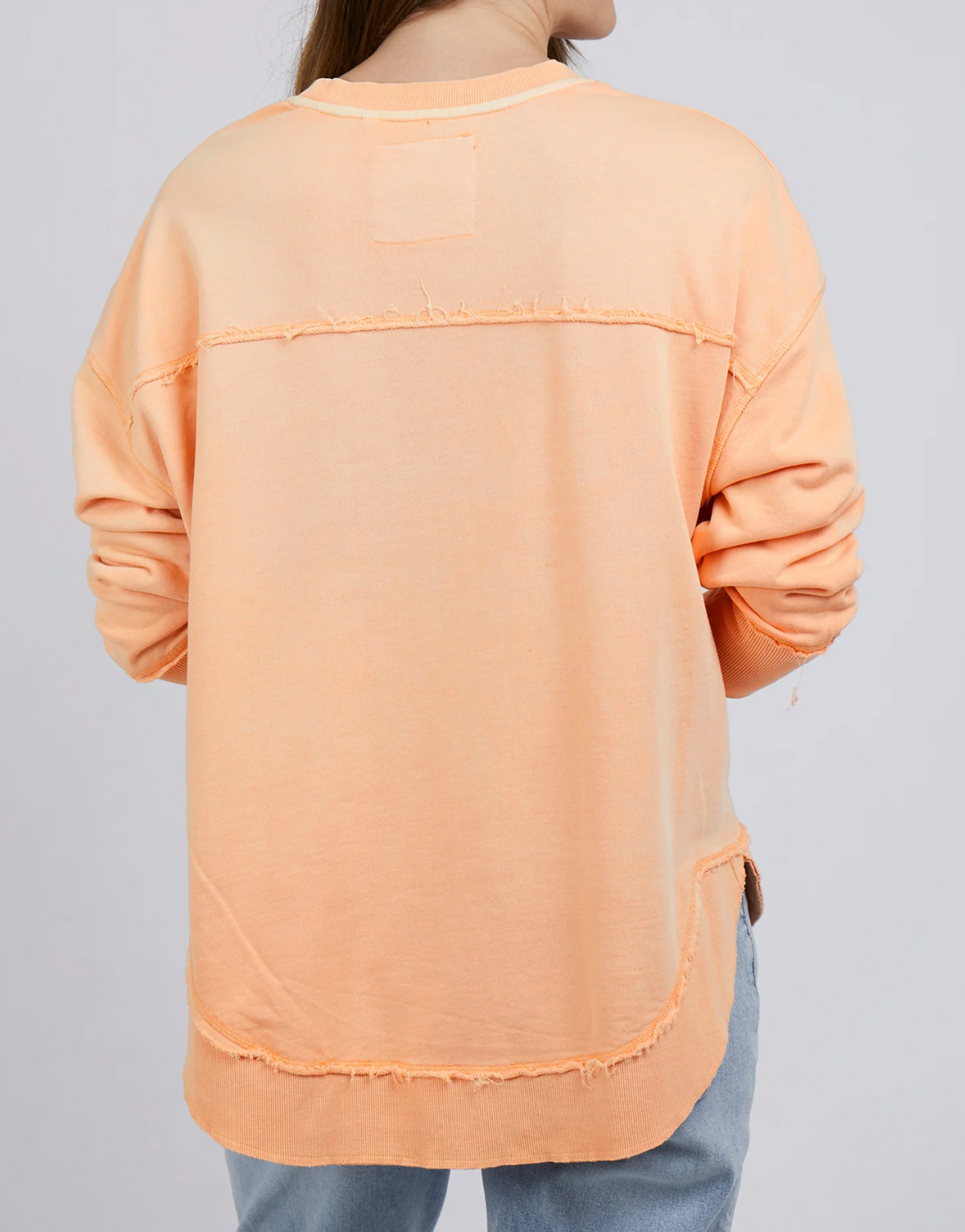 A person wearing a loose-fitting Foxwood Simplified Crew long-sleeve shirt, featuring delicate embroidery and frayed edges in peach color, stands facing away. They pair it with light blue jeans against a plain gray background.