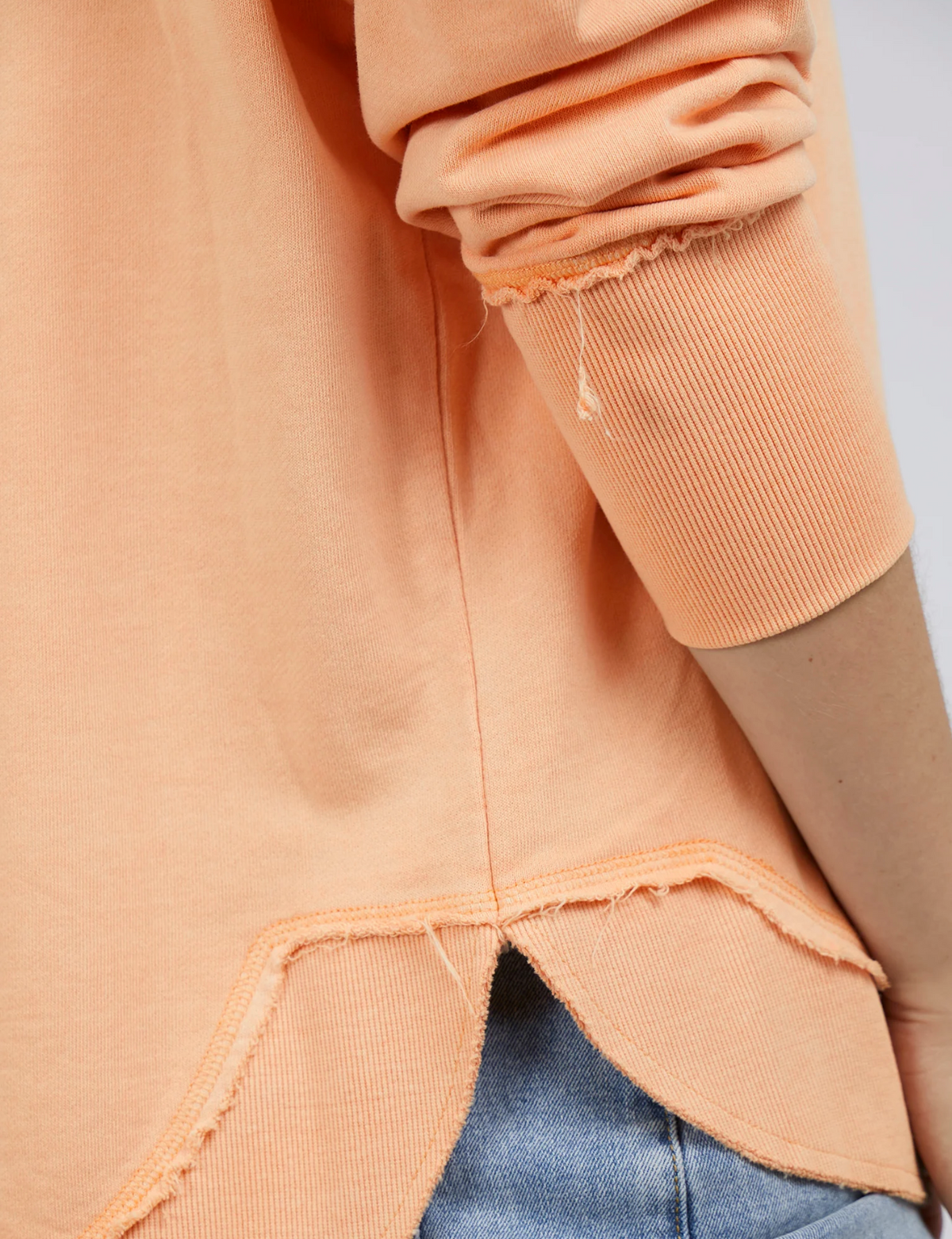 Close-up of a person wearing the Foxwood Simplified Crew, a peach-colored, long-sleeve shirt with embroidery. It features frayed edges, a unique hem design, and is crafted from 100% cotton. Paired with blue jeans, the focus is on its texture and intricate hem details.