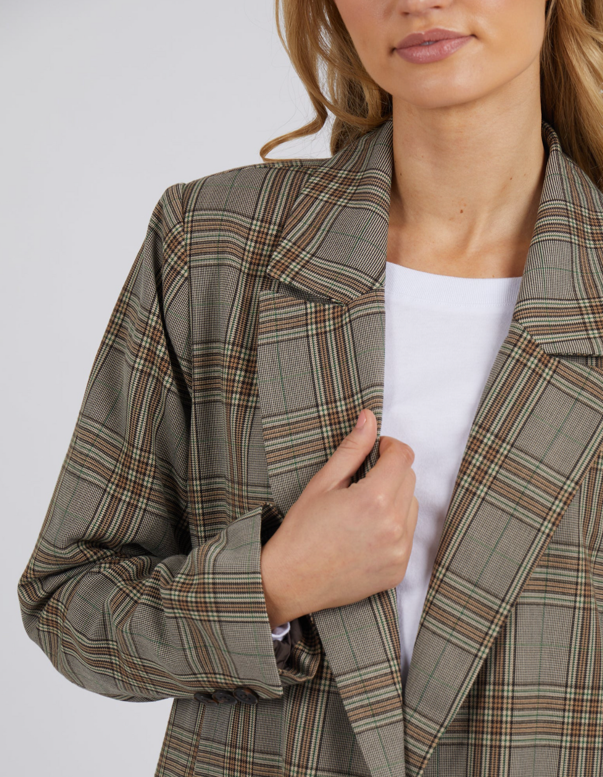 A person in the Foxwood Beatrix Blazer with button details over a white top holds the lapel; their face is partly visible, showing light wavy hair and natural makeup.
