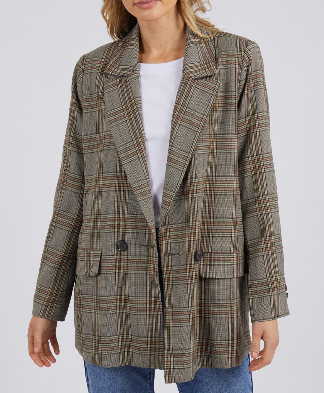 An individual sports the Foxwood Beatrix Blazer, an oversized plaid jacket in brown and gray tones, layered over a white shirt and blue jeans. This comfortable blazer boasts a double-breasted style with dark buttons and flap pockets, set against a plain gray backdrop.