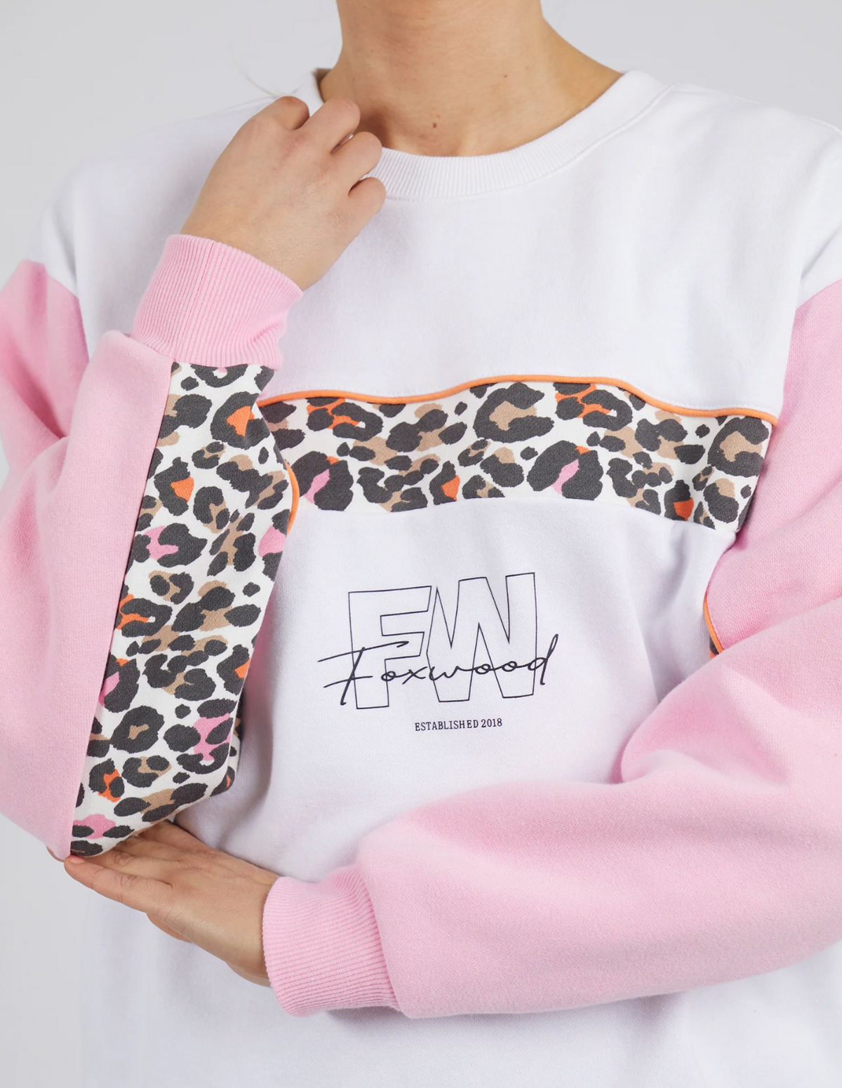 A person in the Foxwood Leopard Crew—100% cotton, with pink sleeves, a leopard print stripe across the chest and sleeves, black text, and a signature-style front graphic—holds their hand near their neck.