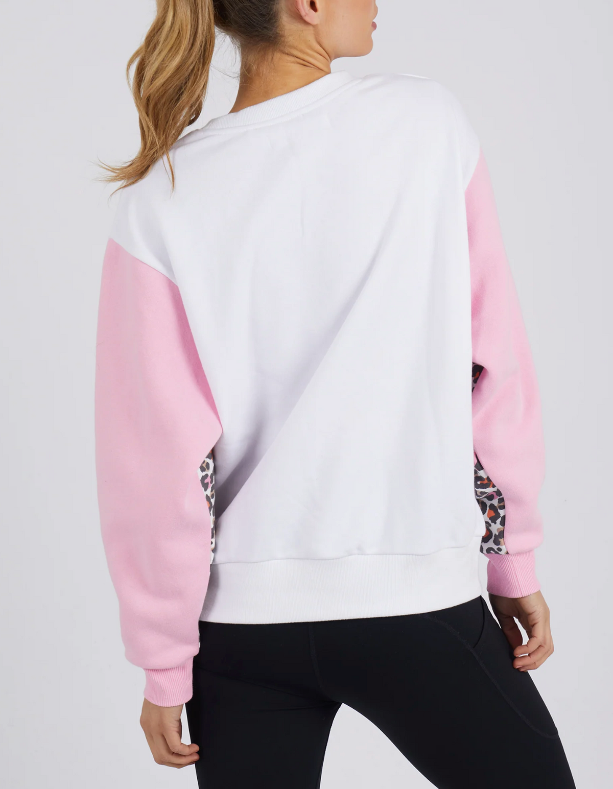 A woman with a ponytail wears the Foxwood Leopard Crew—a white 100% cotton sweatshirt with light pink sleeves and leopard print accents—paired with black leggings. She stands with her back to the camera against a plain background.