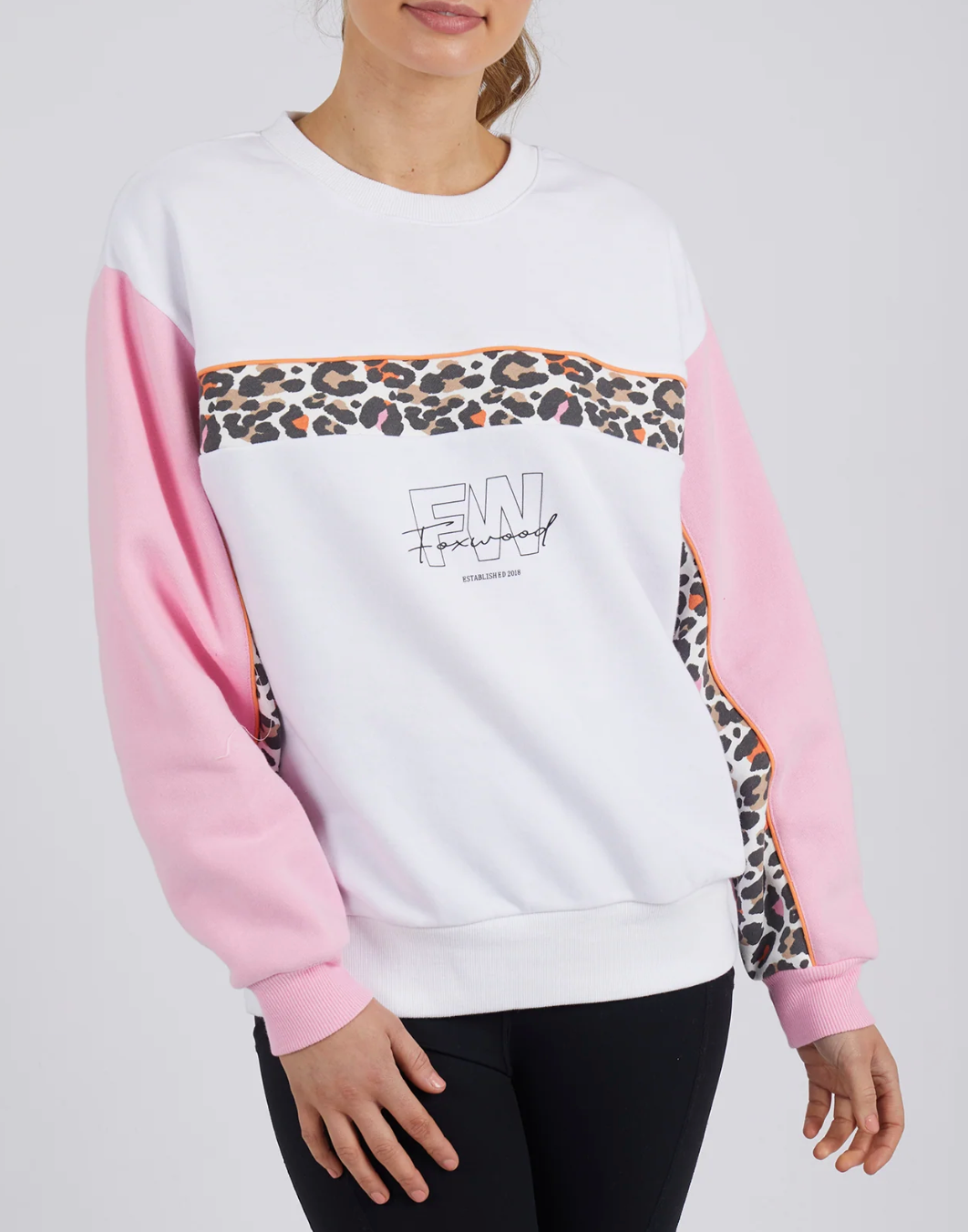 A person wearing the Foxwood Leopard Crew, a white jumper with pink sleeves, leopard print stripe, orange trim, and front graphic text. Made from 100% cotton and styled with black pants.