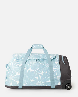 The Rip Curl Jupiter 80L Travel Bag is a large duffel made from recycled polyester, featuring a light blue floral pattern with matching blue handles and wheels. It has a retractable handle and zippered top, set against a plain gray background.