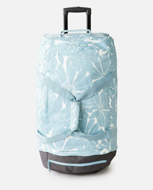 The Rip Curl Jupiter 80L Travel Bag is a stylish, light blue floral-patterned rolling duffel made from recycled polyester. It has a telescopic handle and multiple straps, with a sturdy black base, providing travel convenience and eco-friendly flair.