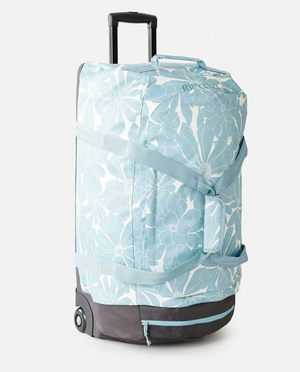 The Rip Curl Jupiter 80L Travel Bag, a light blue and white floral rolling duffel, is made of recycled polyester. It includes black wheels, a sturdy base, a retractable handle, and a side shoulder strap. The top features the iconic brand logo.