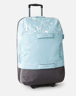 The Rip Curl F-Light Global 110L Sessions Travel Bag is a light blue and gray rolling suitcase with a floral pattern, made from recycled polyester, featuring an extendable handle and two wheels, shown on a white background.