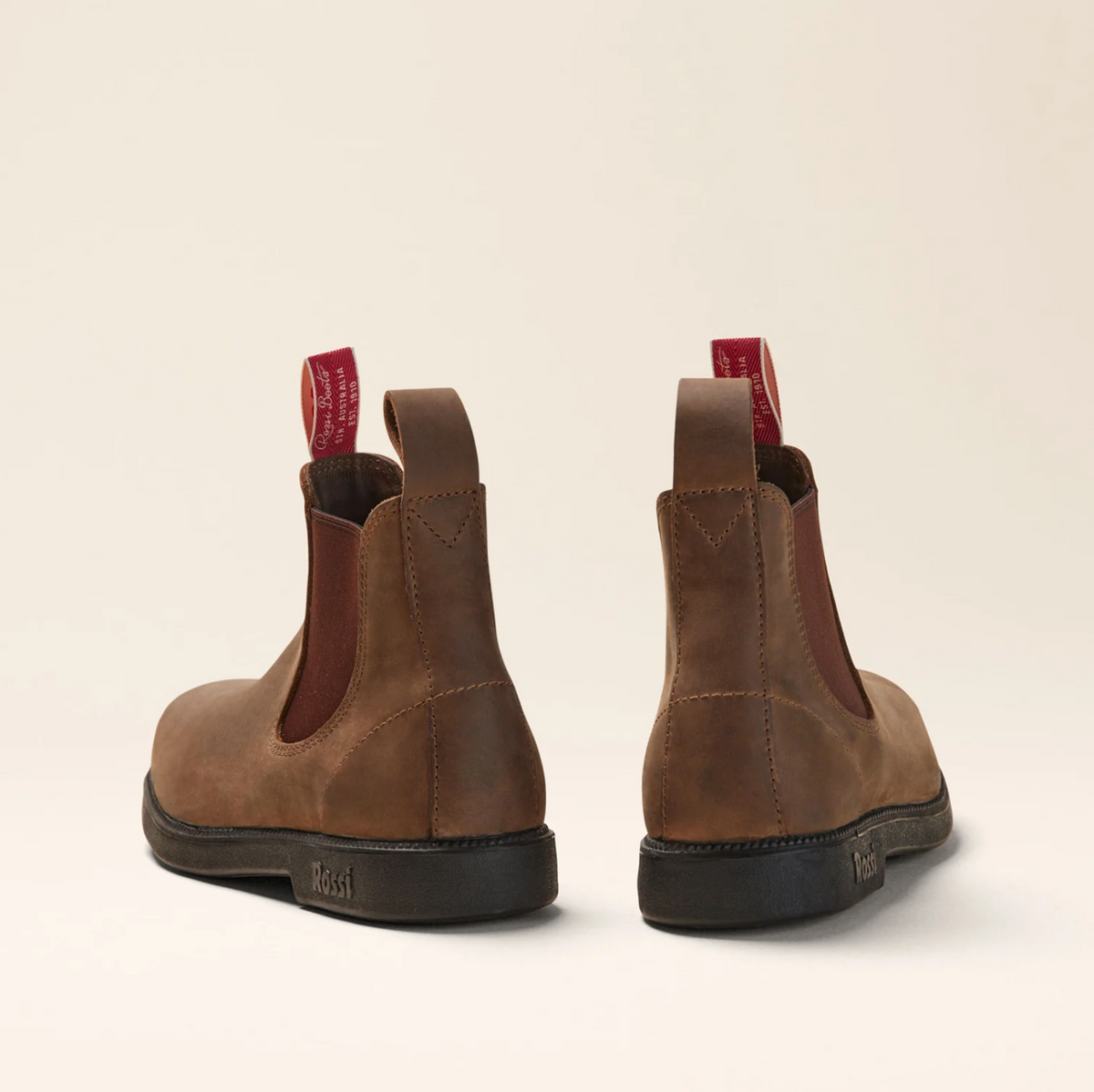 The Rossi Booma 607 Elastic Sided Boot, a pair of brown full grain leather Chelsea boots with black, slip-resistant soles, viewed from the back. The boots feature brown elastic side panels and red pull-tabs at the heel with white text. The background is a plain off-white color.
