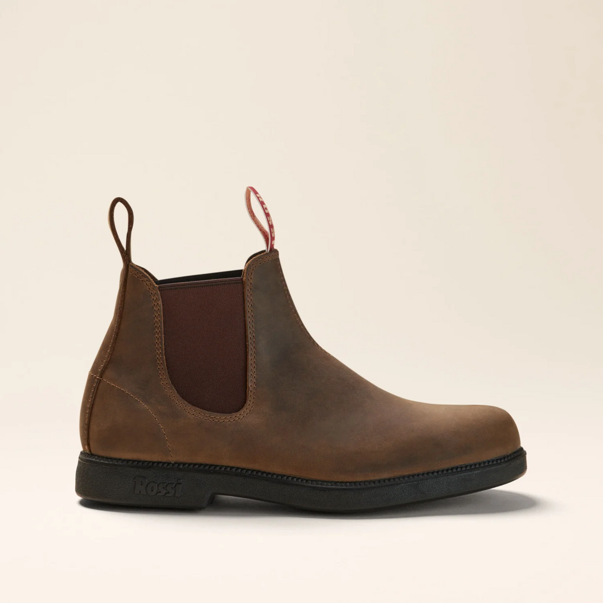 The Rossi Booma 607 Elastic Sided Boot is a single brown slip-on Chelsea boot made from full grain leather, featuring dark brown elastic side panels and pull tabs at the front and back. The boot has a black, slip-resistant sole with the "Rossi" brand name embossed near the heel. The background is a plain beige.