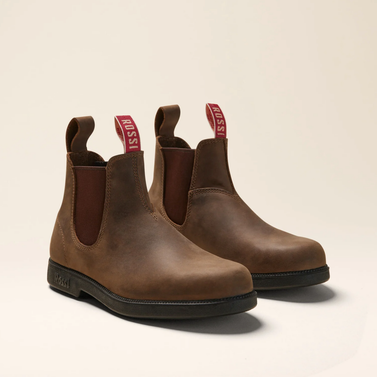 A pair of Rossi Booma 607 Elastic Sided Boots crafted from brown full-grain leather, featuring dark brown elastic side panels and red pull tabs showcasing the Rossi brand. These slip-resistant work boots are displayed on a neutral background.
