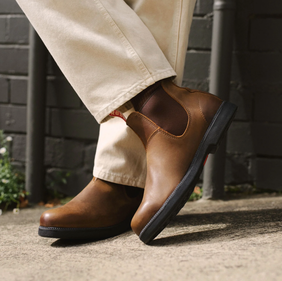 A person wearing beige pants is standing outdoors, showcasing a pair of Rossi Booma 607 Elastic Sided Boots made from Full Grain Australian Leather. The boots feature dark elastic side panels and sturdy black, slip-resistant soles. The surrounding area includes a brick wall and some greenery.