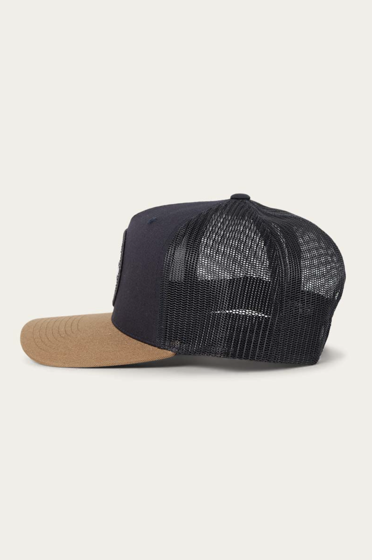 The Ringers Western Signature Bull Trucker Cap features a tan cotton twill curved brim, navy front panel, and black mesh back, displayed in a side view against a plain background.