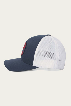 The Ringers Western Signature Bull Trucker Cap is depicted in profile against a plain background. It features a solid blue cotton twill front with a white mesh back and a curved brim, adorned with a circular red patch on the front.