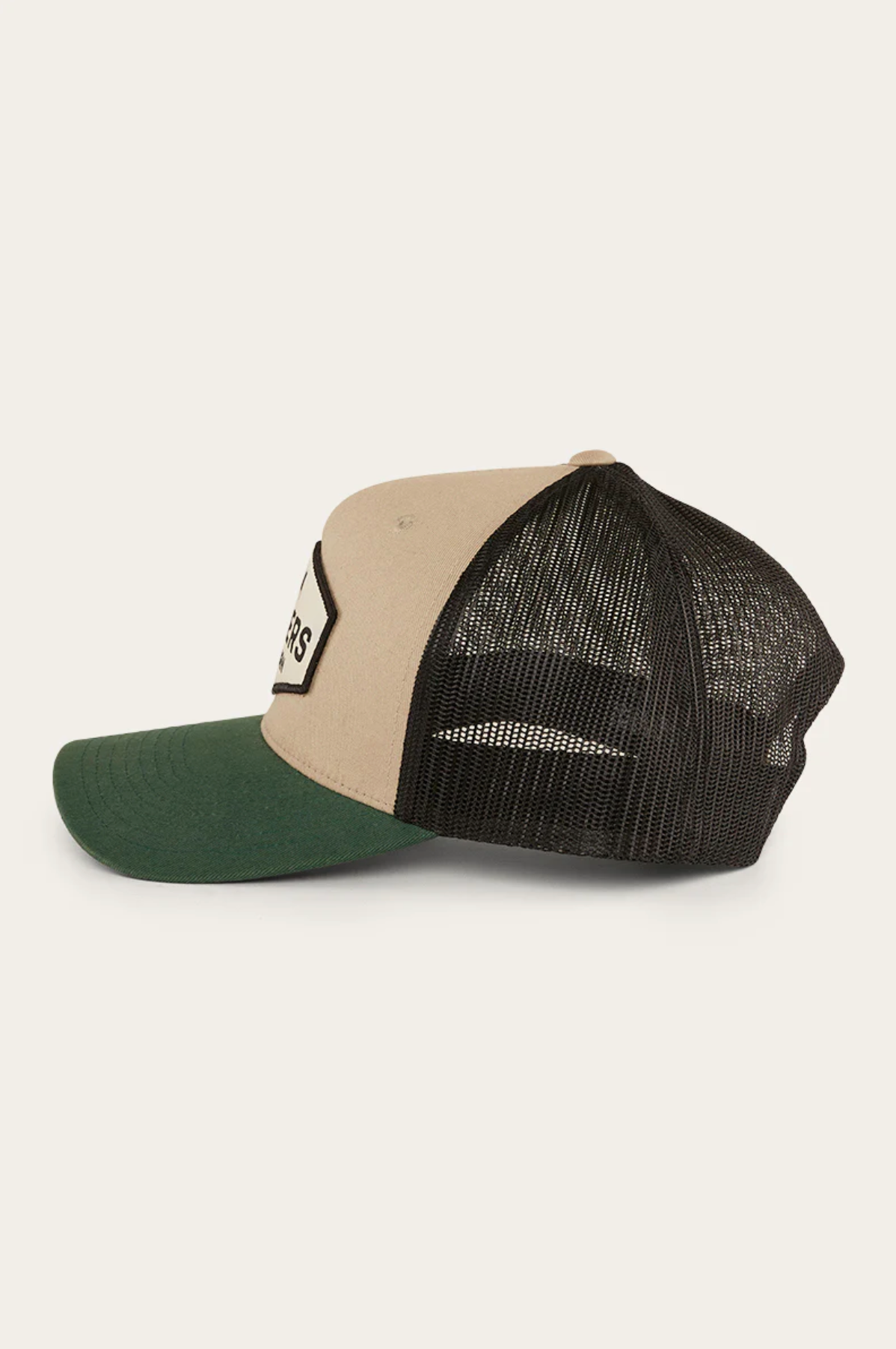 The Ringers Western Scotty Trucker Cap is shown in side view with a beige cotton twill front panel, green brim, and black mesh back. A partial word patch adorns the front. Its classic style stands out against a neutral off-white background.