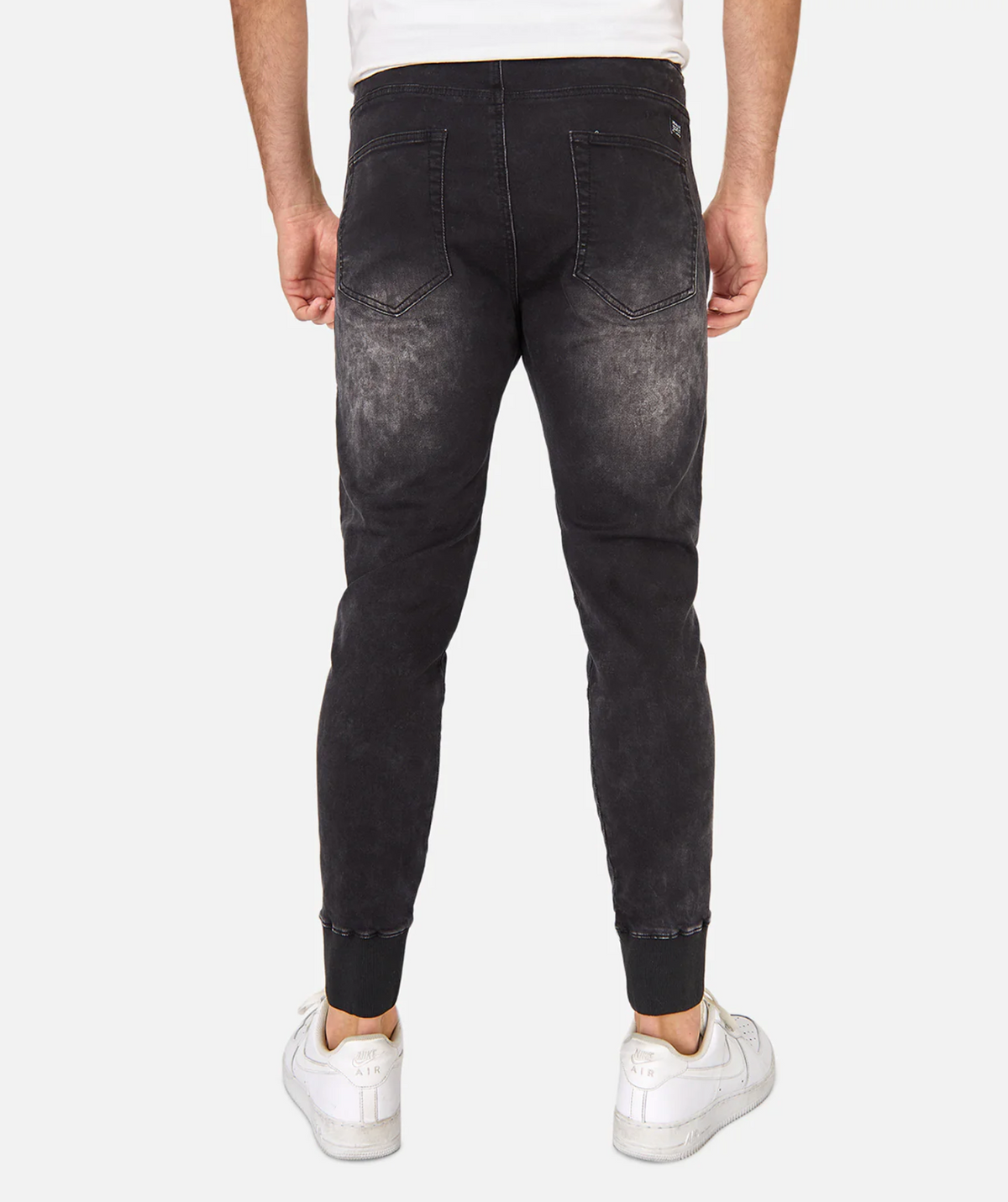 A man faces away, wearing Industrie The Denim Armoured Drifter Pant—jogger-style with an elastic waist and faded back details—paired with white sneakers. The image shows his lower body from the waist down against a plain background.