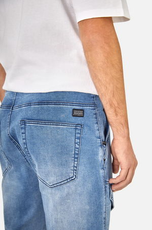 A person is wearing the Industrie The Denim Armoured Drifter Pant, characterized by blue denim and knee panel detailing with a black label on the back pocket, paired with a white t-shirt. The photo is taken from behind, highlighting the stylish pants and capturing the side of their torso and arm.
