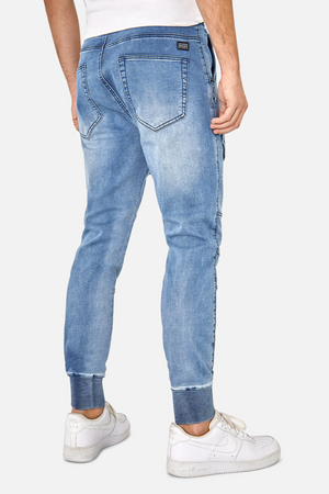 Revised Sentence: Rear view of a person wearing the Industrie The Denim Armoured Drifter Pant in light blue, featuring an elastic waist and cuffs, knee panel detailing, and back pockets with a small black tag on the right pocket. The stylish joggers are paired with a white T-shirt and completed with white sneakers.