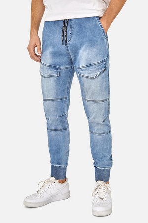 A person is wearing the Industrie The Denim Armoured Drifter Pant in a stylish light blue color, featuring drawstring fastening, multiple pockets, and knee panel detailing. These jogger pants come with an elastic waist for added comfort and are paired with white athletic sneakers and a partially visible white shirt. The background is plain white.