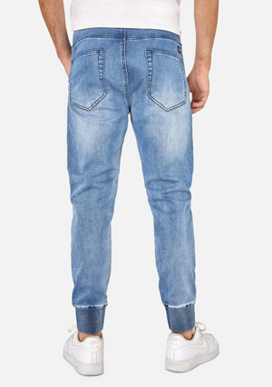 A person wearing Industrie The Denim Armoured Drifter Pant in light blue stands facing away. These denim jeans, highlighted by an elastic waist, feature slight fading on the back pockets and upper thighs. With cuffs folded up to reveal the inner denim, the person completes their outfit with white sneakers and a white t-shirt.
