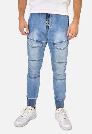 A person wearing stylish Industrie The Denim Armoured Drifter Pant, featuring light blue jogger-style design with knee panel detailing and pockets, stands against a plain white background. They are also wearing a white shirt and white sneakers. The focus is on the lower half of their body.