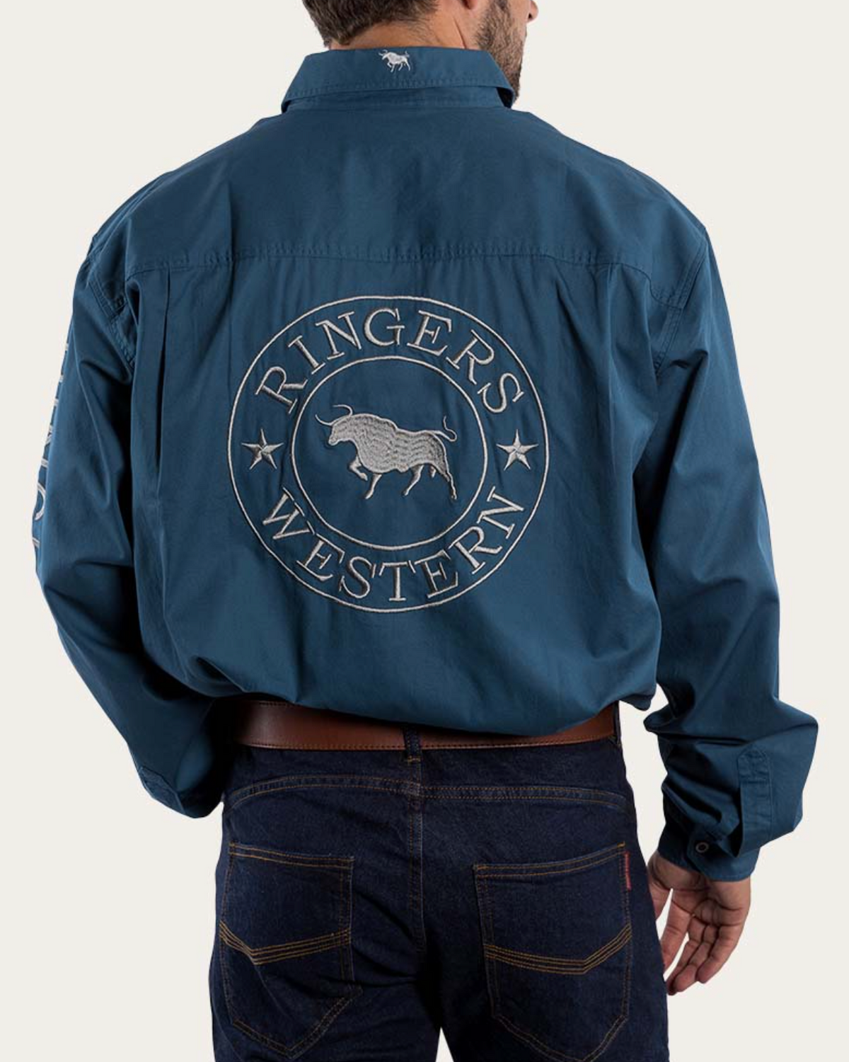 A person wearing the blue Ringers Western Hawkeye Embroidered Shirt with UPF50+, featuring a large circular "Ringers Western" cattle logo on the back and a small logo on the shoulder, paired with dark jeans and a brown belt.