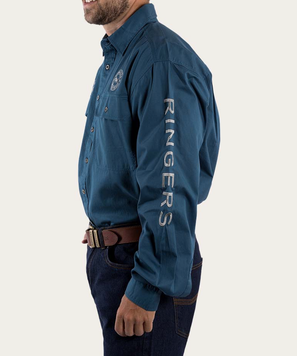 A man sports a Ringers Western Hawkeye Embroidered Shirt, blue with "RINGERS" on the sleeve and an embroidered chest logo, matched with dark jeans and a brown belt. The image crops out his face above the mouth.
