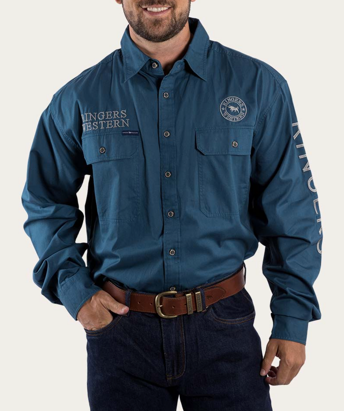 Wearing a Ringers Western Hawkeye Embroidered Shirt with UPF50+ protection, the man sports a blue long-sleeve button-up featuring "Ringers Western" on the sleeve and chest, plus two chest pockets. He pairs it effortlessly with dark jeans and a brown leather belt.