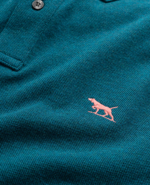 The Rodd & Gunn The Gunn Polo features a teal textured fabric made from premium Australian cotton, with a small pink embroidered dog logo on the right side and partially visible collar and buttons.