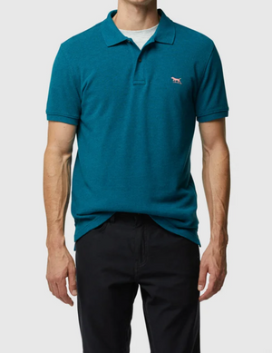 A person wears the Rodd & Gunn The Gunn Polo, a modern-fit teal shirt made from premium Australian cotton with an embroidered emblem on the chest, over a light grey undershirt. The look is paired with sleek black pants against a plain white background.