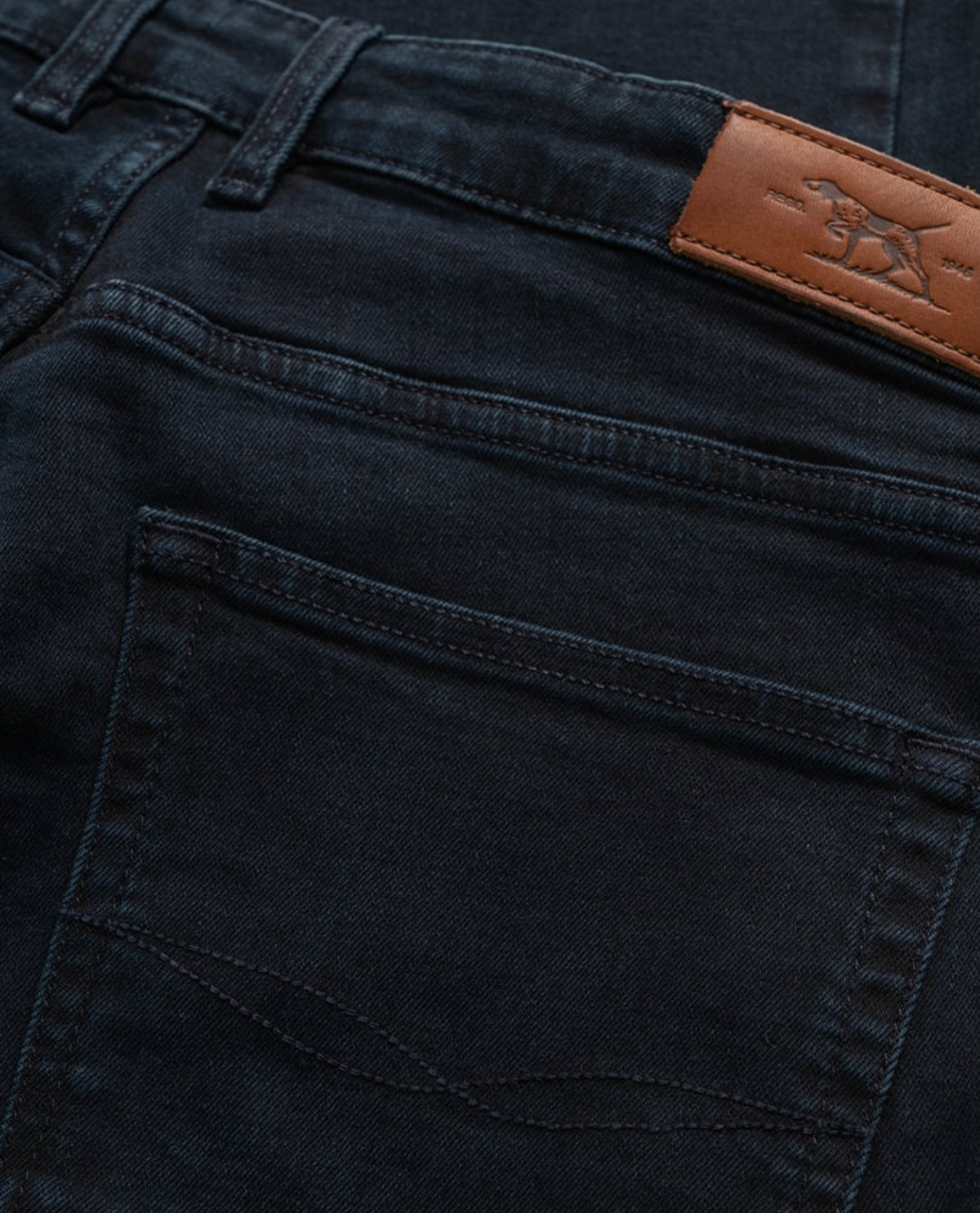 Close-up of Rodd & Gunn's Weston Straight Italian Denim Jeans in deep blue-black, showcasing a visible back pocket. Includes a brown leather patch on the waistband with a horse logo. The comfortable fit, detailed stitching, and fabric texture stand out vividly.
.
