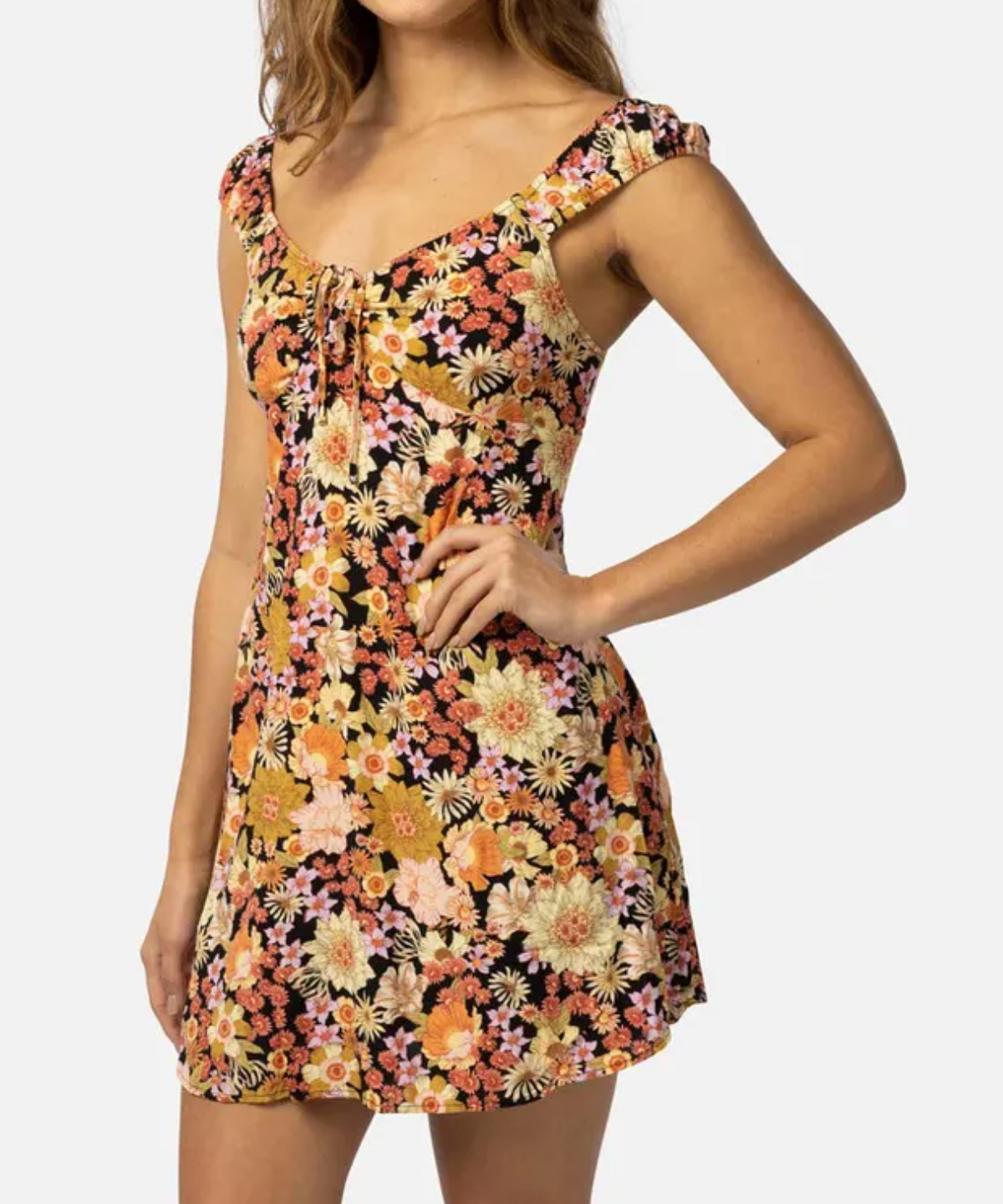The individual is wearing a Rip Curl Mystic Floral Mini Dress made of ECOVERO Viscose Poplin, showcasing a yardage print with pink, orange, and yellow flowers. It features a fitted bodice, flared skirt, and tie detail at the neckline.