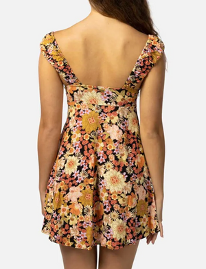 A person with long, wavy brown hair models the Rip Curl Mystic Floral Mini Dress. It features a vibrant print of warm-toned flowers in orange, pink, and yellow, a deep V-back, cap sleeves, and a fitted bodice made from ECOVERO Viscose Poplin against a plain white backdrop.