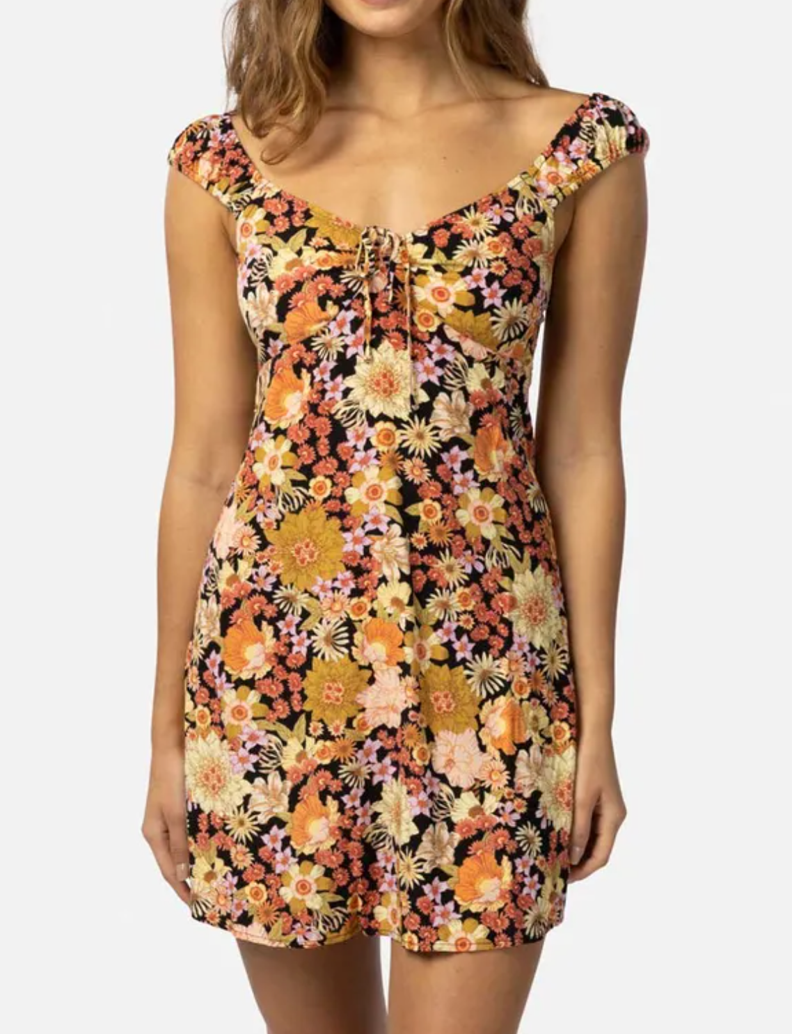 A person models the Rip Curl Mystic Floral Mini Dress, made from ECOVERO Viscose Poplin. The dress features vibrant orange, yellow, and pink floral prints on black, with an off-shoulder design and short sleeves. It is styled against a plain white backdrop.
