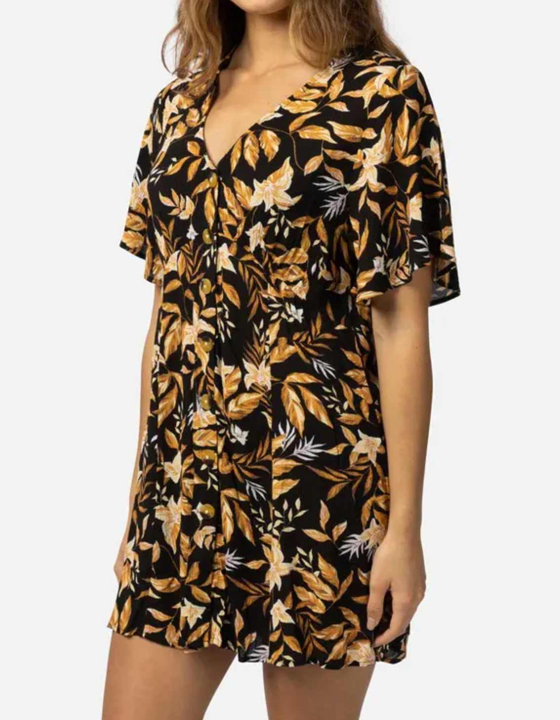 A person wears the Rip Curl La Isla Button Through Dress, crafted from ECOVERO Viscose Crinkle, with a black base adorned with yellow and white floral patterns. This beach-ready dress features short sleeves and a V-neckline against a plain white background.