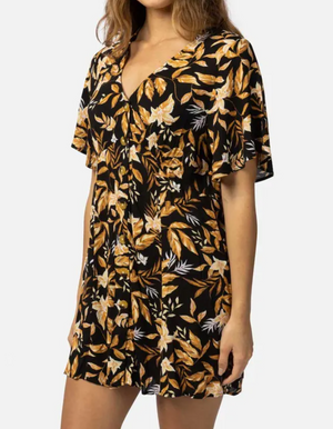 A person wears the Rip Curl La Isla Button Through Dress, crafted from ECOVERO Viscose Crinkle, with a black base adorned with yellow and white floral patterns. This beach-ready dress features short sleeves and a V-neckline against a plain white background.
