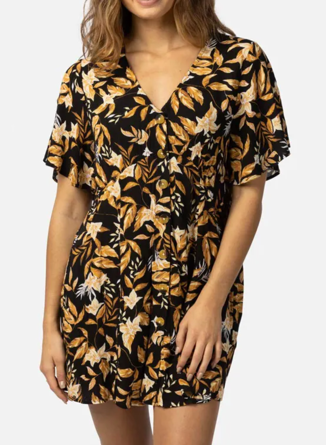 The person is wearing the Rip Curl La Isla Button Through Dress, featuring yellow and white florals on black. Perfect for the beach with its V-neck and short sleeves, this dress is made from ECOVERO Viscose Crinkle. Their wavy hair gracefully falls over their shoulders.