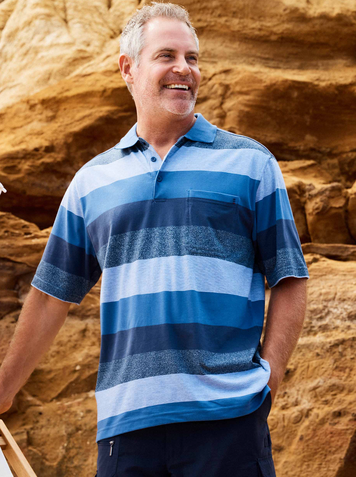 A smiling man with short gray hair wears a Breakaway Neon Kinnersly Polo with a rib knit collar and dark shorts, standing outdoors before large tan rock formations.