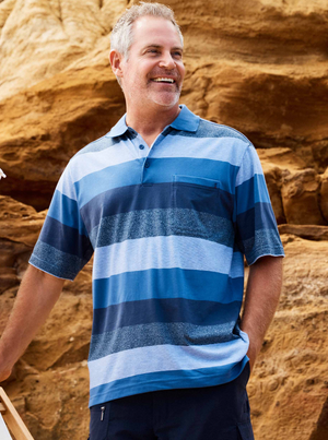 A smiling man with short gray hair wears a Breakaway Neon Kinnersly Polo with a rib knit collar and dark shorts, standing outdoors before large tan rock formations.