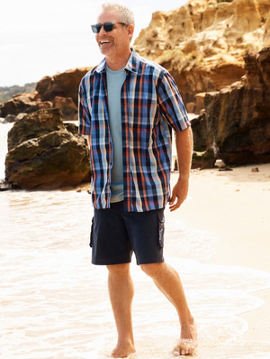 A gray-haired man, in sunglasses and a Breakaway Rome Bamboo Blend Shirt paired with dark shorts, strolls barefoot along a sandy beach backed by rocky cliffs under the sunny sky.
