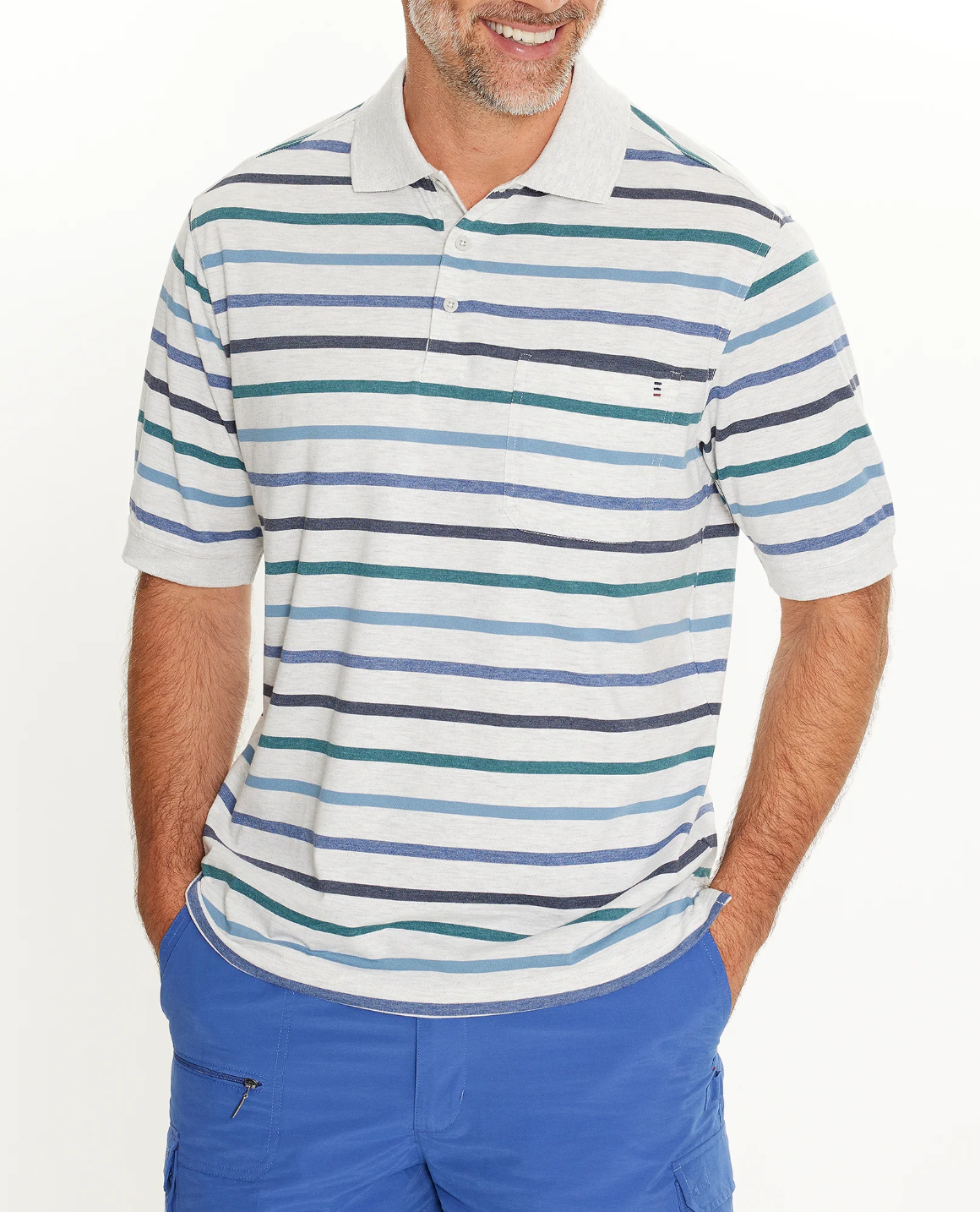 Wearing a Breakaway Ozi Kinnersly Polo with a rib knit collar and white, gray, blue, green, and black accents, paired with bright blue pants, the person smiles warmly with hands in pockets.