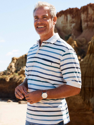A smiling man with gray hair stands on a beach with rugged cliffs, wearing a Breakaway Ozi Kinnersly Polo featuring a rib knit collar. It's a clear, sunny day, and he's holding something in his hand and wearing a watch on his left wrist.